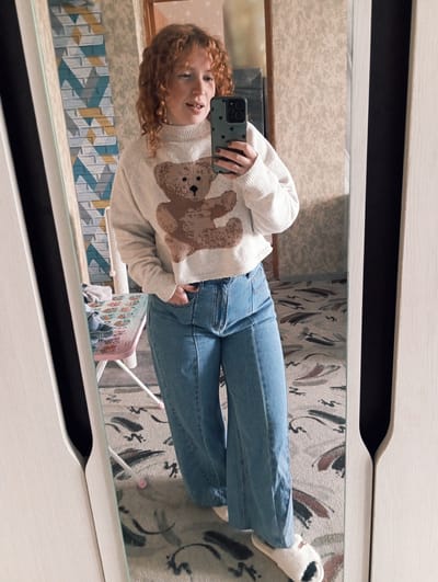 This mirror selfie captures a young individual with shoulder-length, curly red hair, reflected in a large mirror indoors. The person, appearing to be female, is dressed in a casual outfit consisting of a light cream-colored knitted sweater featuring a prominent brown teddy bear design, blue denim jeans, and white furry slippers with a black patch. She holds a smartphone with a dark green, patterned case in her left hand, positioned to take the picture, while her right hand is casually tucked into a jeans pocket. She looks towards the camera with a relaxed, friendly smile.
The background, visible through the mirror, reveals an indoor setting, likely a room in Yurgamysh, Russia. A geometric wall with blue, white, and yellow block patterns is seen to the left. On a surface below, a pink board displaying an alphabet chart with letters (e.g., "Aa Bb Cc", "Rr", "Xx") and numbers (e.g., "1 2 3") is visible, alongside part of a light-colored cat scratcher. The floor is covered by a light-toned rug or carpet featuring abstract, organic designs in shades of brown, black, and grey. The mirror itself is framed by light-colored furniture, possibly a wardrobe or dresser, with a dark handle or recess. The overall scene conveys a comfortable and casual moment.