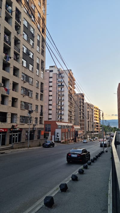 This is a landscape photograph taken in Tbilisi, Georgia, depicting a street scene with several multi-story buildings and a road. The image is oriented vertically, with the buildings dominating the left side and the road on the right.

The buildings appear to be residential or mixed-use, with many windows and balconies visible. Some buildings have a more modern, unfinished appearance, with exposed concrete and scaffolding. Others have a more completed facade, with windows and balconies suggesting habitation. Laundry is visible hanging from some balconies, indicating daily life activities. Utility wires crisscross the sky between the buildings.

On the right side of the image, a road runs alongside the buildings. Several cars are present on the road, including a black sedan and a dark blue vehicle. Black, geometric bollards are spaced along the edge of the road, separating it from the sidewalk.

The sky is a clear, pale blue, suggesting it is either early morning or late afternoon. The lighting indicates a relatively low sun angle, casting soft shadows and bathing the buildings in a warm light, particularly on the right-hand buildings. The overall atmosphere is one of an urban environment during a quiet period of the day. No specific events or activities are clearly depicted beyond the presence of cars and the general urban setting. There is no visible text in the image.
