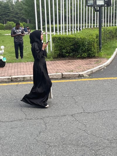 In Changchun City, China, a woman wearing a black hijab and a long black pleated dress walks on an asphalt road with a yellow line marking the edge of a brick path. She is holding a smartphone and a white bag. In the background, a man in a camouflage shirt and black pants stands on the grass, also looking at his phone, with a backpack on. Several people are seated on the grass behind him. A white electric scooter is parked on the left side of the image. The environment includes trees, bushes, and a white fence with vertical poles. A light fixture is visible on a pole behind the fence.