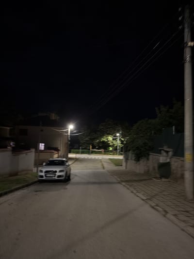 At night, a street in Panagyurishte, Bulgaria, is illuminated by streetlights. A white car is parked on the right side of the street. Power lines are visible above. Vegetation lines the road. Buildings are seen on either side of the street.