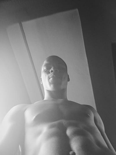A black and white photograph shows a shirtless man with a muscular build, from a low-angle perspective. The man's head is tilted down, facing the camera. The background appears to be an indoor space with a plain wall and ceiling. The location in Nkpor, Nigeria cannot be determined from the image.