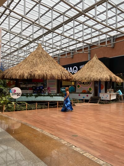 The image captures an indoor market scene in Pampatar, Venezuela, featuring two thatched-roof kiosks set against a modern, grid-like ceiling structure made of metal and translucent panels. The kiosk on the left appears to be a gift shop, indicated by signage that includes "ANUSUAL gifts," "GALOS," and "BOLS." It displays balloons and various items behind a counter. The kiosk on the right, labeled "quipao bubble tea," sells beverages. A woman in a flowing blue dress is walking between the two kiosks, her back mostly to the camera. Another person is seated at the bubble tea kiosk. The flooring is a polished wooden surface with a water feature or reflective area in the foreground. The environment is well-lit, likely from the overhead natural light filtering through the ceiling.