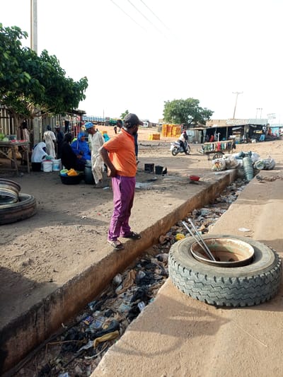 This is a medium shot, outdoor, eye-level photograph of a market scene in Gombe, Nigeria. The foreground features a large, worn truck tire lying on a concrete surface. Behind the tire, a narrow, open drainage ditch is filled with a significant amount of refuse, including plastic bags, bottles, and other debris. A person in an orange t-shirt and purple pants stands to the left of the ditch, facing away from the camera.
In the midground, a group of people are gathered around stalls. Some individuals are dressed in traditional attire, including headscarves and robes. One person is wearing a blue hijab. There are also several buckets and containers visible. A cart loaded with items, possibly goods for sale, is present. Further back, a motorcycle is being ridden by a person wearing a red head covering. The background shows several makeshift market stalls with corrugated metal roofs. In the distance, there are several trucks and a utility pole with wires. The ground is predominantly dirt and sand, indicative of a dry, dusty environment. The overall impression is one of a bustling, informal marketplace with elements of urban decay due to the overflowing drainage.