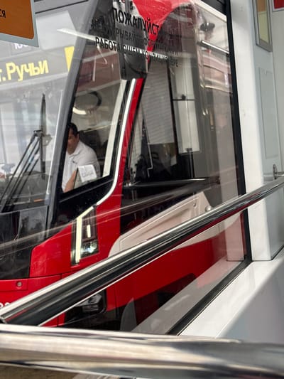 The image shows the interior of a public transport vehicle, possibly a bus or tram, with a driver in the foreground. The driver, a male with dark hair, is wearing a white collared shirt and appears to be looking forward. Reflecting in the window are signs with Russian text, one reading "ПОЖАЛУЙСТА" (Please). Another visible sign in yellow lettering on the side of the vehicle reads "т. Ручьи" which likely refers to the settlement of Ruchey in Sinyavino, Russia. The vehicle's interior is marked by red and white accents. A metal handrail is visible in the lower part of the frame, suggesting it is a passenger area. The scene is captured from a slightly elevated perspective, looking into the driver's cabin.