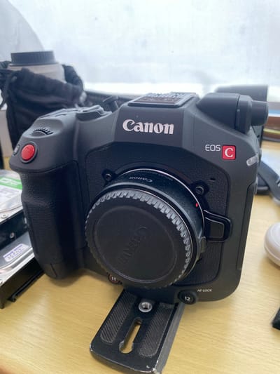 The image shows a black Canon EOS C camera with a lens cap attached, resting on a wooden surface. The camera is positioned on a black tripod mount. To the left of the camera, partially visible, is a hard drive with a sticker. In the background, a black bag with a white lens is discernible. The overall scene is an indoor setting, likely a room with a window showing an overcast sky. The city of Astana, Kazakhstan, is not depicted or implied in the image.