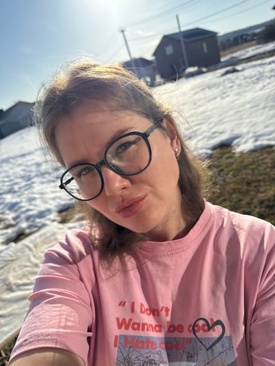A woman is taking a selfie outdoors in Gornyi, Dvurechenskoe S/P, Russia. She is wearing round glasses and a pink t-shirt with a print. A building is visible in the background, along with a patch of snow-covered ground mixed with grass. The sun is shining brightly.
