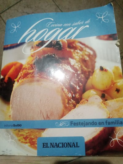 This image is a photograph of a cookbook titled "Cocina con sabor de hogar." The cover features a close-up image of a roasted meat dish with potatoes and what appear to be dried fruits. The text "Cocina con sabor de hogar" is prominently displayed in stylized white font on a blue background. Below the main image, there are two blue rectangular bars. The left bar contains the text "editorialSol90" in white, and the right bar contains the text "Festejando en familia" in white. At the bottom of the cookbook cover, another blue bar displays the text "EL NACIONAL" in white. The background of the photograph is a tiled surface, suggesting the cookbook is placed on a floor or a table with a tiled pattern. There is no discernible location in the city of Santa Ana, Venezuela, as the image is of a cookbook cover and does not depict any specific place or landmarks from that city.