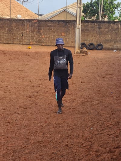 The image displays a man standing on reddish-brown earth under bright daylight. The man is positioned slightly to the left of the frame's center, facing towards the viewer with a neutral expression. He wears a dark long-sleeved shirt, dark shorts featuring a light green band with "SURF UP" printed on it, and dark sandals. On his head is a blue bucket hat. A grey sling bag with the words "Naked" and "22.11.2023" visible is worn across his chest.
The setting appears to be an outdoor, open area. Behind the man, a low wall constructed from reddish-brown bricks stretches horizontally across the mid-ground. Above and behind this wall, lush green foliage from trees is visible, along with parts of a light-colored building roof and a chain-link fence. A concrete utility pole stands vertically on the left side, close to the man. To the right of the man and against the brick wall, three dark tires are stacked. The ground, which is the dominant feature of the lower and right parts of the image, is textured with footprints and scattered small debris, consistent with laterite soil. The bright, clear lighting suggests it is daytime, likely sunny. This environment, characterized by its reddish soil, brick structures, and vegetation, aligns with the location context of Barakin Lerawi, Nigeria.