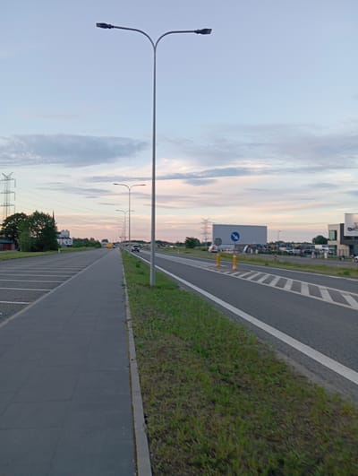 The image depicts a sidewalk next to a road in Dybów Kolonia, Poland. The sky is overcast with soft hues of blue and pink. On the left side of the image, there is a long, empty parking lot with clearly marked spaces. A sidewalk runs along the edge of the parking lot, paved with dark grey rectangular tiles. To the right of the sidewalk is a strip of green grass separating it from the road. The road itself is asphalt and has a white line marking its edge. Further to the right, there are multiple lanes of traffic and what appears to be a parking area with several vehicles. In the background, there are trees, a building that resembles a church with a tall steeple, and power lines supported by large metal towers. Several streetlights, characteristic of modern urban or suburban areas, are visible along the road. A large, rectangular billboard is present in the distance, accompanied by traffic cones and road signage. The scene appears calm and is captured during either dawn or dusk.