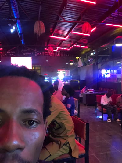This is a medium-shot, eye-level, indoor, low-angle photograph of a group of people in what appears to be a dimly lit bar or lounge. The scene is illuminated by a combination of neon lights, primarily red and blue, that cast colored reflections on the subjects and the environment.
In the foreground, the left half of a Black male's face is prominently featured, taking up a significant portion of the image. His left eye is visible, with a dark pupil and iris, and his skin appears to be reflecting the ambient red light. His dark, curly hair is also visible at the top of his head.
In the mid-ground, other individuals are seated and standing. To the right of the foreground subject, a person wearing a yellow garment is partially visible, and behind them, another person in a blue hooded garment can be seen. Further in the background, several other people are seated on chairs and sofas, some appearing to be looking at their phones. The furniture includes dark-colored chairs and sofas with red upholstery.
The environment is characterized by a corrugated metal ceiling with numerous red neon lights running along its length. Hanging from the ceiling are decorative, spherical objects that appear to be made of straw or similar material. In the background, there are shelves with various items and what looks like a bar area with a television screen or digital display. The floor is dark and reflective, further contributing to the low-light atmosphere.
The overall impression is that of a lively but dimly lit social gathering space, possibly a bar or entertainment venue, with patrons enjoying themselves under the vibrant neon lighting. The location is stated to be Rukuba Road, Nigeria.