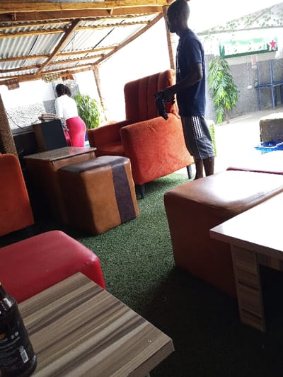 This image captures a semi-outdoor lounge area in the city of Adonkolo, Nigeria. The scene features a corrugated metal roof supported by wooden beams and a green artificial turf floor. Two individuals are present: a male standing in profile on the right, wearing a dark t-shirt and striped shorts, holding a dark object in his hands; and a female in the background on the left, facing away from the camera, dressed in a white top and red skirt, positioned near a light-colored counter. The area is furnished with multiple orange upholstered armchairs, brown and red upholstered ottomans, and small wooden tables. A dark bottle, possibly a stout, is visible on a table in the foreground. In the background, a green leafy plant and a sign with green lettering and a red star are discernible. The scene is brightly illuminated by natural light.