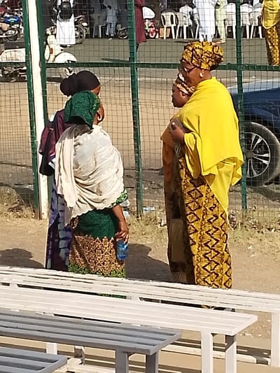 The scene is located in Sabon Gari East, Nigeria. Three adult females and one child are positioned in the midground, partially obscured by white slatted benches in the foreground.

The woman on the far left, facing away from the viewer, wears a dark head covering, a purple and blue patterned garment, a green head wrap, and a light-colored shawl. She holds a blue plastic water bottle in her right hand. Behind her, a second woman is partially visible, also wearing a dark head covering and purple/blue patterned clothing. The third woman, on the right, faces left and wears a yellow headwrap, sunglasses, gold-colored earrings, a yellow top, and a yellow and black patterned long skirt. Her left arm is around a child positioned in front of her, who wears a similar patterned garment and head covering.

A metal chain-link fence, with green and dark red posts, separates the subjects from the background. Behind the fence, a white scooter is visible on the left. To the right, part of a dark-colored vehicle is visible. Numerous indistinct figures, some seated on white plastic chairs, are present in the distant background, suggesting a public gathering. The ground is earthen, and shadows indicate outdoor daylight conditions.