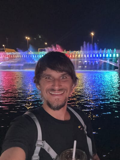 A medium shot captures a man holding a beverage in front of a colorfully lit fountain. The man has short, dark hair, a light complexion, and a light beard. He wears a black shirt with grey straps across his shoulders. The fountain displays various colors from top to bottom, including blue, green, yellow, orange, and red. The location is Wulingyuan, China.