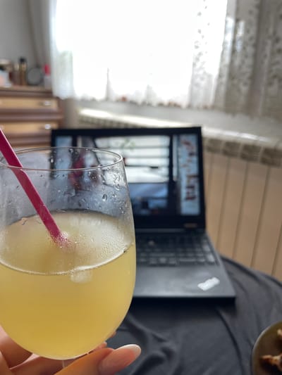 A hand holding a glass of yellow liquid with a pink straw is in the foreground. The glass is sweating, indicating it is cold. In the background, a laptop is open on a dark cloth surface. Behind the laptop, a wooden dresser with drawers is visible, and a white lace curtain hangs over a window. The overall scene appears to be an indoor setting, possibly a home or office, in the city of Zlatograd, Bulgaria.