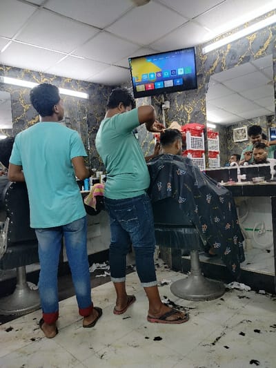 The image shows the interior of a barbershop in Dhaka, Bangladesh, located at the coordinates 23.784 latitude and 90.377 longitude. A man in a turquoise shirt is standing to the left of the frame, while another man in a teal shirt is cutting the hair of a customer seated in a barber's chair. The customer has a black cape around his shoulders. The barbershop is dimly lit and has a flat-screen TV playing on the wall. The floor is covered in tiles and there are black hair clippings scattered around. There is a large mirror on the wall behind the customer, reflecting a man in a white shirt who appears to be having his hair cut. There is another man in a blue shirt standing to the right of the mirror. All of the men are wearing flip-flops.