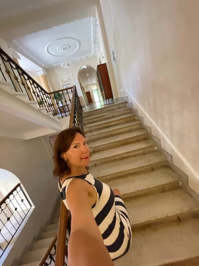 In Gagra, Abkhazia, a woman with brown hair and red lipstick, wearing a striped dress, takes a selfie on a grand staircase. The staircase features stone steps and ornate railings. The background reveals a high-ceilinged hallway with intricate plasterwork on the ceiling, a chandelier, and doorways leading to other parts of the building. The overall impression is of an elegant, classical interior.
