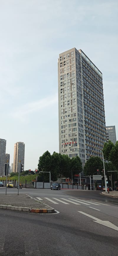 The image shows a street scene in Jiulongpo District, Chongqing, China. A tall, rectangular building dominates the left side of the frame. It has numerous windows in a grid pattern and appears to be an apartment or office building. Adjacent to the building are trees. Traffic lights with a red signal are visible. A black SUV is parked on the side of the road. A street with an arrow pointing to the right and a crosswalk is on the right. In the background, more buildings can be seen, as well as a bit of green space. The sky is overcast.