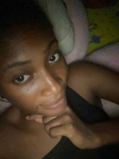 A dark-skinned individual is lying down, with their head resting on a pillow. They are looking directly at the camera with a neutral expression. The lighting in the room is dim, creating a shadowy atmosphere. The background consists of a blurred green and pink patterned surface, possibly a blanket or a rug. The image appears to have been taken indoors, likely in a bedroom. There are no visible landmarks or contextual clues to indicate the specific location within Kubwa, Nigeria. The subject is wearing a dark colored top.