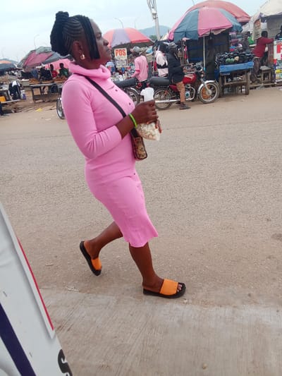 In Kubwa, Nigeria, a woman in a pink, form-fitting, long-sleeved dress with a high neckline walks across a paved surface. Her hair is styled in braids with short, blunt bangs. She wears large gold hoop earrings and orange and black slide sandals. A leopard-print purse hangs across her body, and she holds a clear plastic bag filled with what appears to be popcorn in her right hand. The background depicts an outdoor market scene with various stalls and umbrellas, a motorcycle parked nearby, and other people visible among the vendors and customers. The overall atmosphere suggests a bustling marketplace.