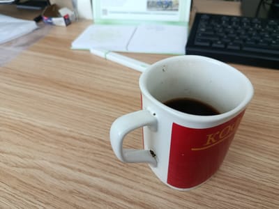 The image depicts a close-up view of a white coffee mug with a red band and yellow text on a wooden desk. The mug contains dark liquid, likely coffee. A white pen with a green band is visible in the background, along with a black keyboard and some papers. The scene appears to be an office or workspace. The text on the mug is partially obscured but appears to be in Cyrillic script. The overall impression is one of a casual work environment. The image does not contain any information about the city of Arkadag, Turkmenistan.