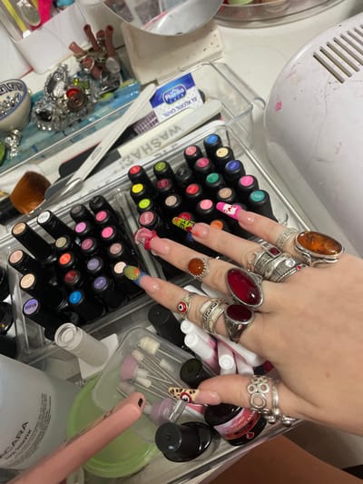 A close-up shows a hand adorned with numerous rings, displaying creative nail art. The hand rests amidst a collection of nail polish bottles arranged in rows, along with various manicure tools, including files, drill bits, and other supplies. Labels like "ACARA," "XELJAYI," "Phorma," and various numbers are visible. The arrangement suggests a well-organized and personalized nail care station.