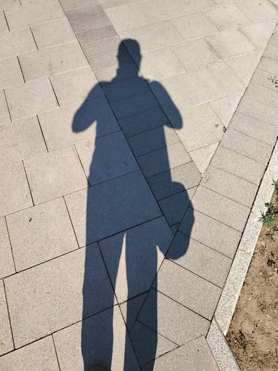 A shadow of a person, cast from the right, falls across a paved surface composed of rectangular stone slabs. The shadow is elongated and shows the outline of a head, torso, arms, and legs. One arm appears to be bent, and the person is holding an object, possibly a bag, which is also visible as a shadow. The paving stones are light gray with dark speckles and are laid in a regular pattern. To the right of the paved area, there is a strip of exposed earth with some dry grass and small plants. The light source creating the shadow is to the right and slightly above the subject, indicating it is daytime. This scene could be located in a public area of Astana, Kazakhstan, such as a park, square, or promenade, given the typical urban landscaping and paving materials found in the city.
