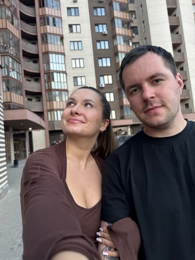 A man and a woman are taking a selfie. The man is wearing a black t-shirt. The woman is wearing a brown long-sleeved top. The backdrop is a multi-story building with numerous windows, located in Odintsovo, Russia.