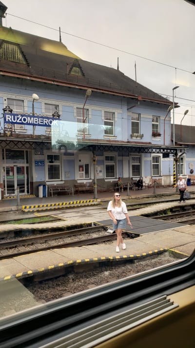The image shows a train station in Ružomberok, Slovakia. The station building is light blue with a dark roof and features multiple windows and awnings. A large blue sign above the entrance reads "RUŽOMBEROK". Several people are visible on the platform. In the foreground, a young woman in a white top and denim shorts walks towards the right. Further back, a woman sits on a bench and an older woman walks with a bag. The train tracks and platform are clearly visible, with yellow and black safety markings along the edge of the platform. The scene appears to be captured from a train window.