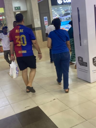 A group of people appear to be gathered inside a shopping mall, possibly waiting in line or observing a display. One person is wearing a Barcelona soccer jersey with the number 20 and the name "ALXE" on the back. Another person is wearing a blue shirt and jeans. The location seems to be a retail area with storefronts and display merchandise visible in the background. The floor is tiled, and the overall setting appears to be a typical shopping mall environment.