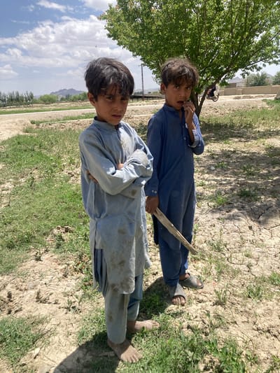 Two boys are lying on their sides on the ground. One boy wears a blue garment and sandals. He holds a stick. The other boy wears a dirtier blue garment. He is barefoot with his arms crossed. The ground is grassy with patches of dirt and cracks. A tree is on the left. Buildings, mountains, and a cloudy sky are in the background. A motorcycle is parked near the tree. The location is outdoors in the city of Qilla Saifullah, Pakistan.