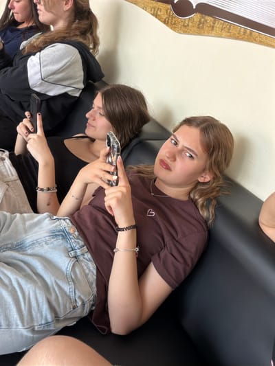 Three young women are seated indoors on a black sofa. The image is taken from a slightly elevated, angled perspective.
The central figure, a young woman with light brown, wavy hair, is looking directly at the camera with a skeptical or annoyed expression, her eyebrows furrowed and lips slightly pursed. She is wearing a brown t-shirt with a small pink heart embroidered on the chest, and light blue denim jeans. She holds a smartphone in her left hand, pointing it with her right index finger towards the woman seated below her. She also wears a silver necklace and multiple bracelets on her wrists.
Below her, another young woman with dark brown hair is visible from the chest up. She is engrossed in her own smartphone, looking down at the screen. She wears a black tank top and light-colored pants, along with several bracelets.
On the far left, a third young woman is partially visible. She has light brown hair and is wearing a black and grey hoodie, also looking downwards, presumably at a device or activity not fully captured.
The background consists of a plain light-colored wall. To the far left, a decorative element, possibly a frame or wall art, features brown and white stripes with some indistinct dark patterns or text on a light brown background. The overall atmosphere suggests a casual, indoor setting, where the individuals are relaxing and engaged with their mobile phones. No specific location cues are visible beyond the provided context of Panagyurishte, Bulgaria.
