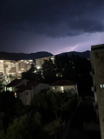 This is a vertical, slightly blurry nighttime photo taken from a high vantage point, possibly a balcony or window, overlooking a city landscape during a thunderstorm. The sky is dark and dominated by heavy, dark purple clouds. A dramatic lightning strike flashes across the upper left portion of the frame, illuminating the clouds with a bright white light.
Below the stormy sky, the silhouette of a mountain range is visible. In the foreground and middle ground, several buildings are illuminated. One prominent building has a facade with many horizontal lines of warm yellow lights, suggesting it is an apartment building or hotel. Other buildings are less distinct, with some windows showing light. Trees with dark foliage are scattered throughout the scene, adding depth and a sense of natural elements within the urban environment. The overall atmosphere is one of a powerful natural event occurring over a city at night, with a sense of awe and perhaps a touch of drama. The location is identified as Bar, Montenegro.