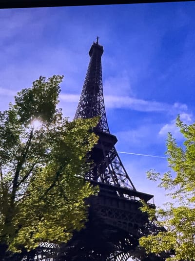 The image shows a low-angle view of the Eiffel Tower against a bright blue sky with some wispy clouds. Lush green trees frame the lower portion of the tower, with sunlight filtering through the leaves. A faint contrail from an airplane is visible in the sky behind the tower. The Eiffel Tower is a wrought-iron lattice tower on the Champ de Mars in Paris, France.