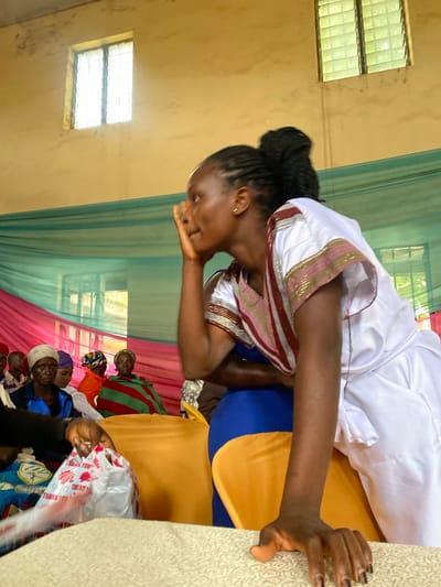 In Riyom, Nigeria, a woman with braided hair pulled into a bun, wearing a white robe with gold and red trim, leans forward with her hand raised to her face, suggesting deep thought or listening. She is positioned in the foreground, facing right, while a group of other women are visible in the background, seated and facing forward. The environment appears to be an indoor gathering, possibly a church or community hall, with pastel-colored drapery adorning the walls and windows with louvered panes. The lighting is bright, with sunlight streaming through the windows. Several attendees are wearing headscarves in various colors and patterns. In the lower left corner, a plastic bag with red lettering reading "THANK YOU" is being held by an unseen person.