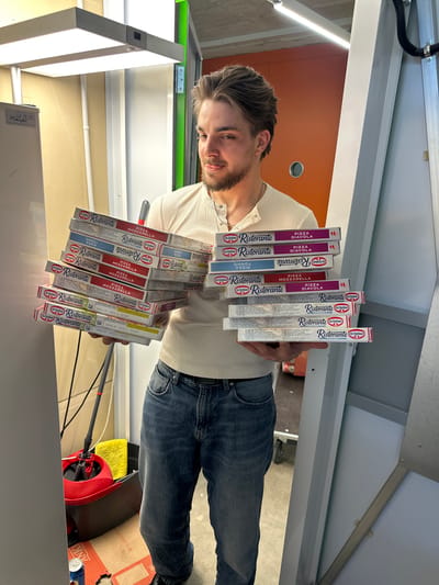 A young man is standing indoors, holding a large stack of Dr. Oetker Ristorante pizza boxes. The boxes are stacked unevenly and feature different pizza flavors including Diavola, Mozzarella, and Tonno. The background reveals an interior space with fluorescent lighting, orange and white walls, and various cleaning supplies, including a mop and bucket. There is a sticker reading 'Mayer' on a white box. Based on the product packaging written in German, and the license plate on the white box, this image could be taken in Volketswil, Switzerland.