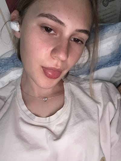 A young female with light brown hair is lying down and sticking her tongue out. She is wearing white earbuds in her left ear and a light-colored t-shirt with a heart-shaped pendant necklace. She is in a room with blue and white striped bedding in the background. There is no information or visual indication of the location being in Panagyurishte, Bulgaria.