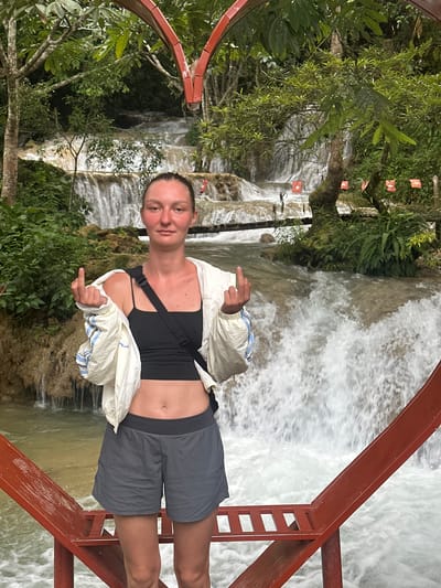 A woman stands on a red metal platform in front of a waterfall in a lush, green environment. She is wearing a black tank top, a white jacket with blue and white striped accents, and grey shorts. Her hair is pulled back in a bun. She is giving a middle finger gesture with both hands. Small red flags with white stars are visible on a tree branch behind her. The waterfall appears to be tiered, with water cascading down multiple levels. The overall scene suggests a tourist location. The location in the city of Cổ Lũng Commune, Vietnam is not evident from the image itself, but the context of lush vegetation and tiered waterfalls is consistent with such locations.