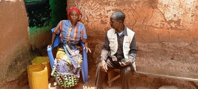 In Makungu, Democratic Republic of the Congo, a woman and a man are situated outdoors against an earthy-toned wall. The woman, wearing a red headwrap, a striped shirt, and a patterned skirt, sits on a blue plastic chair, gesturing with her right hand while speaking. To her right, a man, wearing a dark shirt, trousers, and a light-colored multi-pocketed vest with visible text "UNGSU" (possibly "UNICEF" or similar organization abbreviation), sits on a low wooden stool, holding a small dark object, possibly a voice recorder, in his left hand. He is looking at the woman. Two yellow plastic containers, cut open, are on the ground near the woman's chair. A narrow wooden bench is positioned behind the man. The background consists of a rough, cracked, reddish-brown earthen wall, with a section behind the woman displaying peeling dark green and black paint. The ground is bare earth.