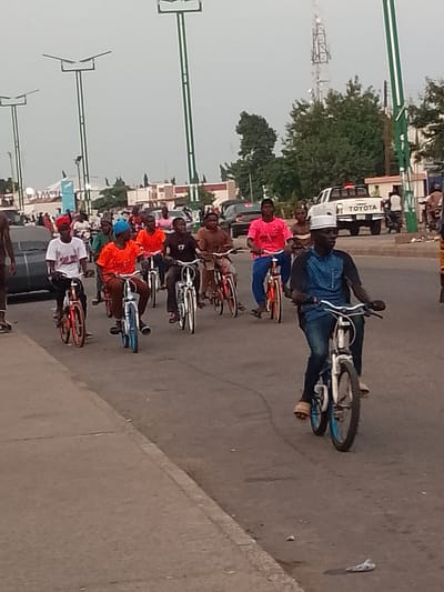 A group of young men and boys are riding bicycles in a procession on a paved road in Pantame, Nigeria. The riders are of varying ages and are dressed in casual clothing, including t-shirts, shirts, and trousers. Some of the cyclists are wearing hats or head coverings. They are riding on the right side of the road, and there are vehicles and streetlights visible in the background. The sky is overcast, suggesting a cloudy day. One man in the foreground, wearing a blue shirt and dark pants, is riding a bicycle with a white frame and blue rims. He is wearing a white cap. Behind him, other cyclists are also visible, some on bicycles with orange rims and others on bicycles with blue rims. A white Toyota pickup truck is parked on the right side of the road in the background. Streetlights with solar panels are present along the road. The overall impression is a casual gathering or parade of cyclists in an urban setting.