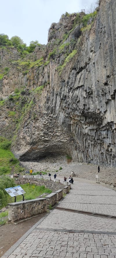 The image depicts the Symphony of the Stones, a natural monument of columnar basalt formations, located near Garni, Armenia. The site is a cliff face composed of hexagonal and pentagonal basalt columns. In the foreground, a stone-paved path leads towards the cliff, with a low stone wall alongside it. Behind the wall is greenery. Several people are present in the image. Some are standing on a small viewing platform near the stone wall, while others are walking along the path towards the cliff face. A sign with text and images stands near the wall. The sky is overcast, suggesting a cloudy day.