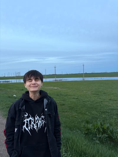 The image shows a person with short dark hair, smiling with eyes closed, wearing a black hoodie with a white graphic print and a black denim jacket with red details. The person has a tattoo on their neck.
The background consists of a sky with light blue hues, a green field, and electrical power lines. Azyakul', Russia, has experienced flooding; the water is visible between the green fields.