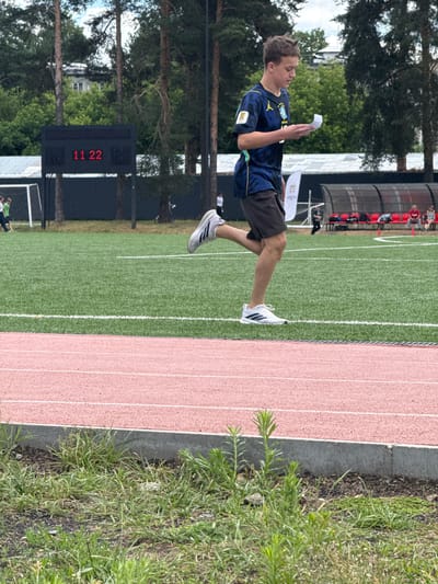 The image depicts a young male athlete in a dark blue and yellow sports jersey and dark shorts, running on a track. He is holding a white piece of paper. The background shows a green sports field with a scoreboard displaying "11 22", trees, bleachers with people, and a soccer goal. The scene appears to be a sports event or training session in Dmitrov, Russia. The foreground is occupied by a red running track and some grass and dirt.