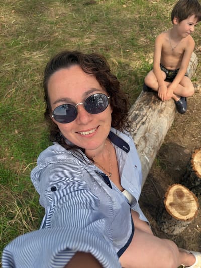 A boy and a woman are outdoors, possibly in a park or a wooded area. The boy is shirtless, wearing dark shorts with a pattern, and has a silver chain around his neck. He is sitting on a large, fallen log, with his legs crossed. To the right of the boy, there are two cut tree stumps. The woman is lying down, in the foreground of the image. She is wearing a blue and white striped button-down shirt, with a dark collar and dark buttons. She is also wearing sunglasses with round, dark lenses and a thin silver frame. A thin gold necklace is visible around her neck. The ground around them is covered in dry grass and some patches of green grass. The overall lighting suggests it is daytime, with bright sunlight. The scene appears relaxed and casual, possibly a family outing. There is a small red tag with white lettering on the woman's shirt, near the collar, which is too blurry to read.