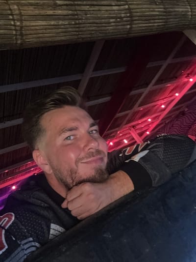 A Caucasian male with short blond hair, light stubble, and light eyes is posing for a photo. He's wearing a dark, patterned sports jersey with white and red accents. His right hand is near his neck. The background features a bamboo-like structure on the left, and a building with a red-lit framework. The image appears to be taken in Canggu, Indonesia.