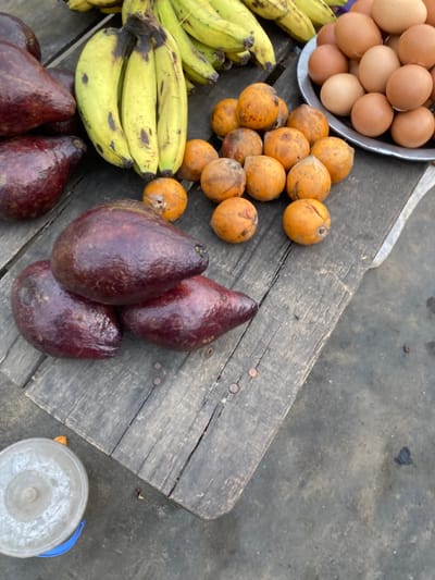A wooden table displays various food items. A silver bowl contains brown eggs. Bunches of yellow bananas lie adjacent to small, round, orange fruits. Four dark, ripe avocados are arranged on the table's surface. A light gray bucket is placed near the table's edge. The backdrop consists of a light gray concrete surface.