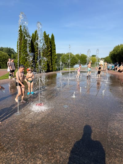 This media file depicts a sunny day in a park in Minsk, Belarus, where children and some adults are cooling off at a splash pad. The scene is dominated by a large, patterned granite surface with numerous water jets spraying upwards, creating a playful and refreshing atmosphere.
Several children are visible, actively engaging with the water. One young boy in swim trunks lies on his back, enjoying the spray, while another child in a swimsuit lies on their stomach, also being showered by the water. Further back, more children are seen interacting with the fountain, some running through the sprays and others lying down. Adults are present, observing or participating in the activity. Some are seated on a brick ledge in the background, while others stand near the water.
The time of day appears to be late afternoon, suggested by the long shadows cast on the ground, including a prominent shadow of a person on the right side of the frame. The weather is clearly warm and sunny, indicated by the clear blue sky and the people's attire, which includes swimsuits and light clothing. Lush green trees and bushes frame the splash pad area, suggesting a well-maintained park environment. The overall mood is one of joy and relaxation, with the splashing water providing a welcome respite from the heat. There is no visible text within the image.