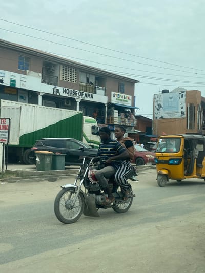 The image captures a daytime street scene in Ado, Nigeria, characterized by urban activity and various commercial establishments. The sky is overcast.

In the foreground, two men are riding a motorcycle on a paved road. The man in front wears a striped blue and white shirt, and the passenger behind him wears a black shirt. Their expressions are neutral, focused on their journey. To their right, part of a yellow auto-rickshaw, identifiable by the "TVS" logo on its front, is visible, suggesting it's also in traffic. Behind the motorcycle, a dark-colored SUV or car is parked or slowly moving. Further down the street, a white truck with green accents can be seen.

The street is lined with multi-story buildings, mostly made of concrete. Many of these buildings house businesses, indicated by numerous signs. Visible text includes "HOUSE OF AMA WHERE WOMEN RULE," "STEPH & VAL KITCHEN WORLD," and a billboard advertising "DON-JOEL SOLAR." Another building displays logos for electronics brands like "Infinix," "oppo," and "Xiaomi," along with the text "LAPTOPS." A sign for "EYE CLINIC CLEVELAND OPTICAL" with a phone number "08055095960" is also present. Overhead power lines crisscross the sky. A large green shipping container is situated near one of the buildings. The overall scene depicts a bustling commercial street environment.
