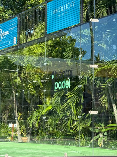 The image shows a padel tennis court enclosed by glass walls, located in Baan Tai, Thailand. Lush green palm trees and other tropical foliage are visible through the glass, creating a backdrop of natural greenery. On the upper part of the enclosure, three blue banners are displayed. The leftmost banner is for "Reform Studio" and includes text about "private classes available for all levels" and "underground pilates." The middle banner is for "MMOLUCKY REAL ESTATE." The rightmost banner is partially visible and appears to be advertising a real estate service as well. On the glass wall itself, a logo with the text "abentoi padel" is visible in green lettering. The court's surface is a bright green synthetic turf. The scene is brightly lit, suggesting it is daytime, and the overall impression is of an outdoor sports facility integrated into a tropical environment. There are no people visible in the image.