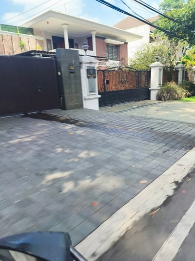 The image shows a paved driveway in front of a house located in Bandung, Indonesia, at the coordinates -6.879 latitude, 107.586 longitude. The driveway is made of gray rectangular tiles and is bordered by a white line. The house is a two-story building with a large front gate and a brown door. There is a white mailbox to the right of the door, and the gate is made of black metal with an ornate design. There is a small patch of green grass and bushes on the right side of the driveway. The driveway is shaded by the house, creating a pattern of light and shadow on the tiles. The street is visible in the foreground, and there is a vehicle's rearview mirror in the lower right corner. The image is taken from the point of view of a person looking down the driveway towards the house.