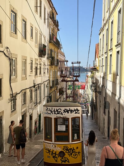 This is a daytime, eye-level, full shot of the iconic Tram 28 climbing a steep, narrow, cobblestone street in Lisbon, Portugal. The tram, a vintage yellow model with graffiti on its side, is the central focus. The street is lined with tall, narrow, light-yellow buildings with white trim around the windows and wrought-iron balconies. Utility wires crisscross the sky.
In the foreground, a woman with blonde hair pulled back in a bun is facing away from the camera, wearing a black tank top with a green strap and a tattoo on her upper back. To her left and slightly further away, a woman with dark hair is wearing a white dress with a polka-dot pattern, also facing away from the camera and gesturing with her hands.
Further up the street, on the left side, a man in a green t-shirt and black shorts with a backpack is facing the camera, and beside him, a woman in a light green dress is facing the man. They are standing on the sidewalk next to the tram tracks.
The street continues uphill, narrowing in perspective, with more buildings on either side and a glimpse of a body of water at the end of the street, possibly the Tagus River. Flags, including a Brazilian flag, are visible hanging from structures above the street. The sky is clear and blue. The overall scene depicts a busy, picturesque street in the Alfama district, known for its historic trams and narrow streets.